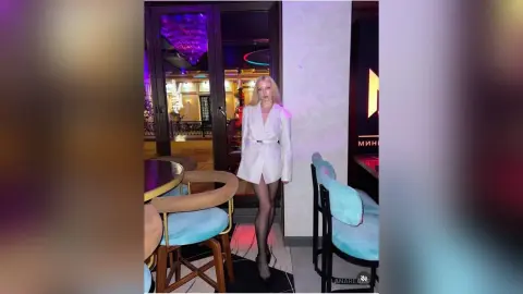 Elegant woman in a white blazer and black tights standing in a modern, dimly lit lounge setting.