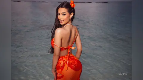 Carine poses on a beach wearing an orange floral bikini, looking back over her shoulder with long dark hair.