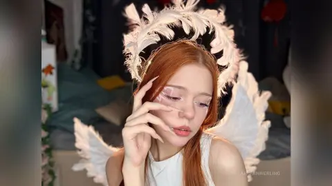 Redheaded woman in white angel costume with feathered wings and ethereal makeup in a cozy bedroom setting.