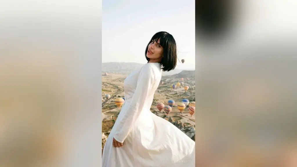 SandraBex — Elegant woman in a flowing white dress overlooking hot air balloons in a dreamy Cappadocia landscape.