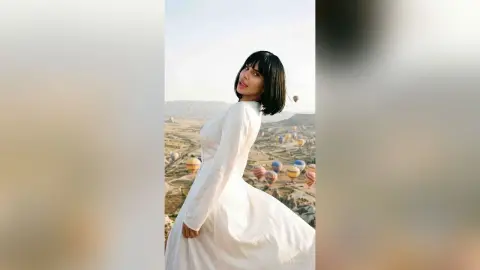 Elegant woman in a flowing white dress overlooking hot air balloons in a dreamy Cappadocia landscape.