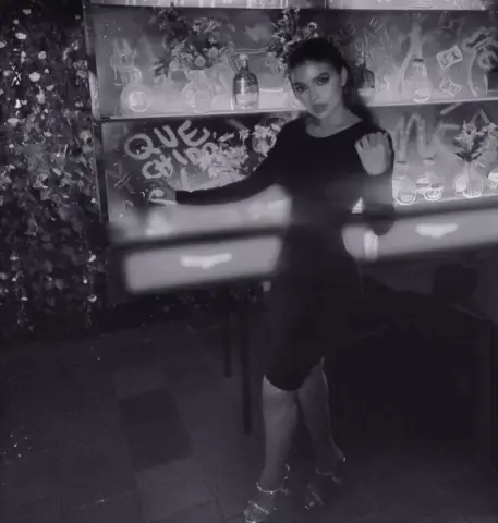 Stylish woman in a black dress posing in a moody, dimly lit bar with floral decor.
