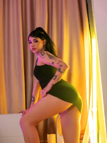 Young woman with tattoos in a green mini dress posing against gold curtains in warm, moody lighting.