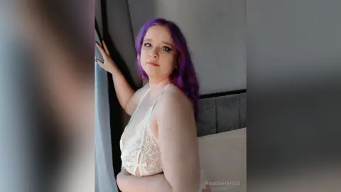 Young woman with purple hair in white lace lingerie posing by a window in a bright bedroom.