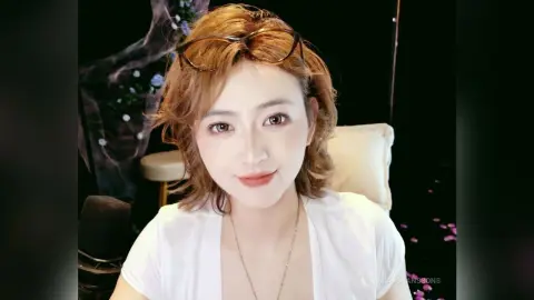 Smiling young woman with wavy brown hair wearing a white top and glasses in a cozy indoor setting.