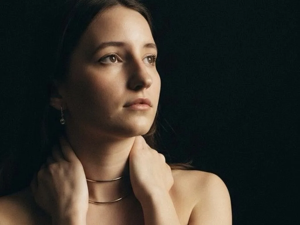 KoriPengra looks off-camera while holding her neck with both hands against a dark background.