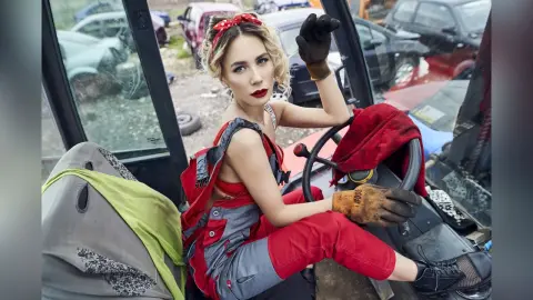 ChristineDavis poses in red and grey overalls and black gloves, sitting in a vehicle surrounded by scrap cars.