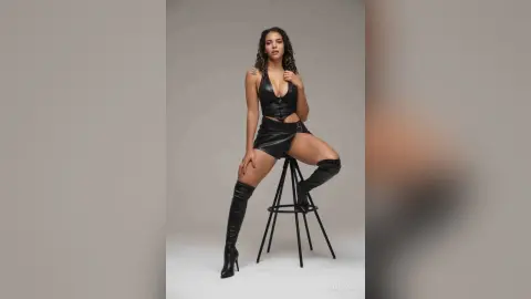 Confident woman in a black leather bodysuit and over-the-knee boots posing on a stool in a studio setting.