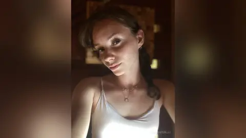 Young woman in a white camisole tank top posing in warm, moody indoor lighting.