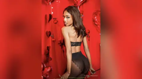 Glamorous woman in black lace lingerie posing against a romantic red heart balloon backdrop.