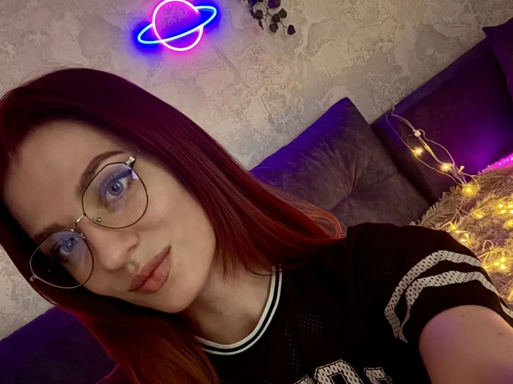 JulietJess poses close-up wearing glasses and a black mesh t-shirt against a purple sofa and neon light.