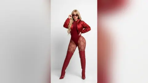 Confident woman in a red bodysuit, quilted jacket, and over-the-knee boots posing against a white background.