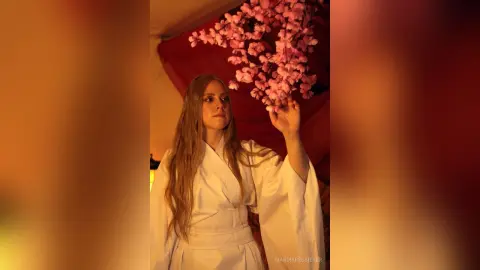 Blonde woman in a white flowing robe standing under pink cherry blossoms in a warm, dreamy setting.