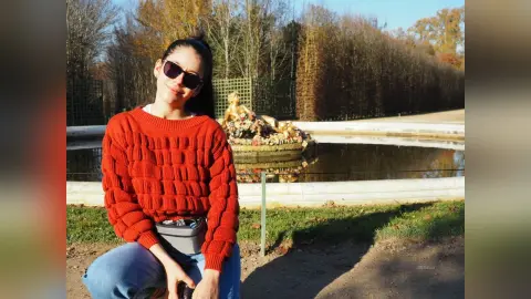 JessieLiu wears a red textured sweater and sunglasses while sitting outdoors near a pond surrounded by trees.