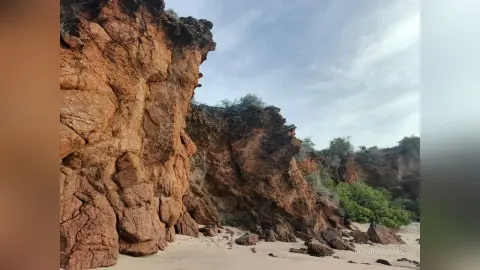 Rugged brown rock cliffs overlooking a sandy beach under a cloudy sky in a natural landscape.