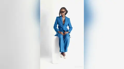 Stylish Black woman in a blue power suit and sunglasses sitting on a white pedestal against a clean background.