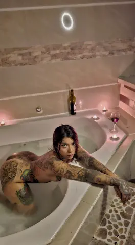 carolinewolftv is a tattooed woman posing naked in a white bathtub with a bottle of wine nearby.