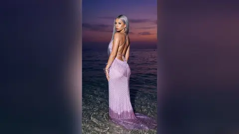 Kalira poses in a long, light purple backless dress while standing in shallow ocean water at sunset.