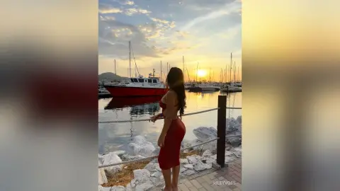 Woman in a red dress standing on a pier overlooking sailboats at sunset in a peaceful marina.