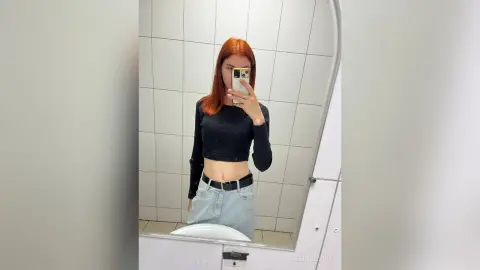 Redheaded woman in a black long-sleeve crop top and light denim jeans taking a mirror selfie in a bathroom.