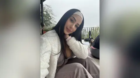 Smiling woman in a white fuzzy cardigan and grey loungewear relaxing on an outdoor balcony.