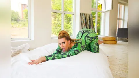 NataliiaSnake licks her lips while posing on all fours on a white bed, wearing a tight green snakeskin print long-sleeve bodysuit.