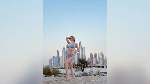 BellaBronze poses in a light blue bikini on a sandy beach with a city skyline and ocean in the background.