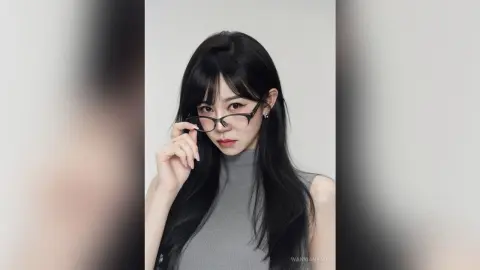 Young woman with long black hair and glasses wearing a grey turtleneck in a minimalist studio setting.