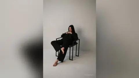 Stylish woman in a black jumpsuit sitting on a chair in a minimalist white studio setting.