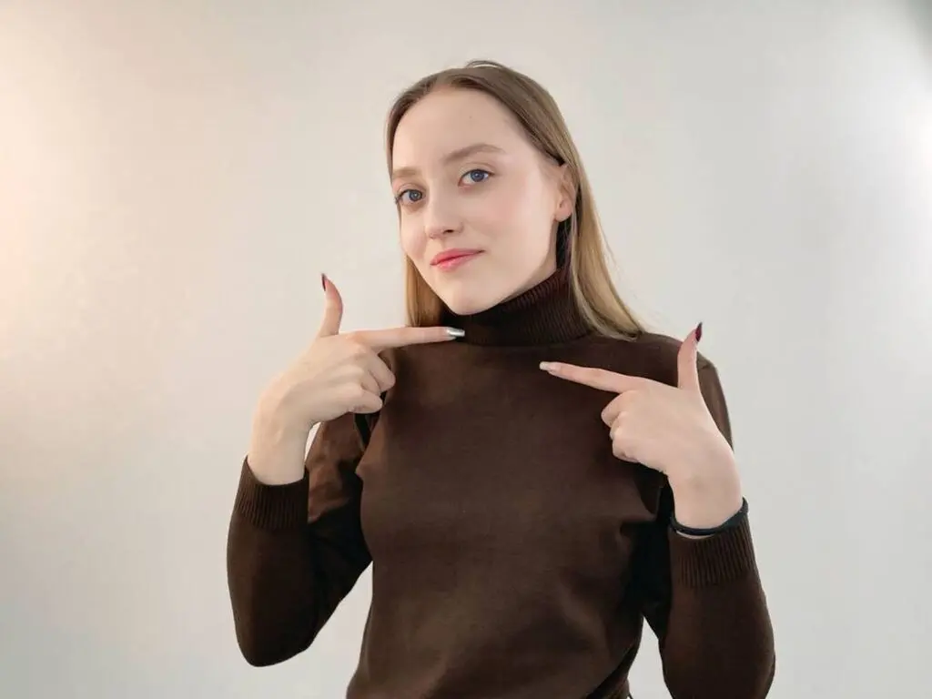 Young woman in a brown turtleneck sweater pointing to her neck against a plain white background.