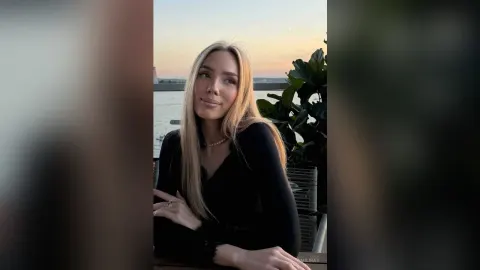 AmiliRay — AmiliRay with long blonde hair wears a black top and pearl necklace while sitting outdoors by the water at sunset.