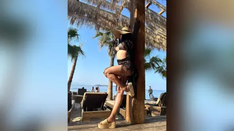 NicoleBella stands outdoors in a black lace bra and matching bottoms, wearing a wide-brimmed straw hat against a sunny beach backdrop.