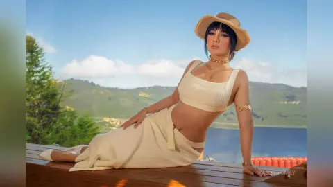 Stylish woman in a white ribbed crop top and skirt wearing a straw hat, relaxing by a scenic mountain lake.