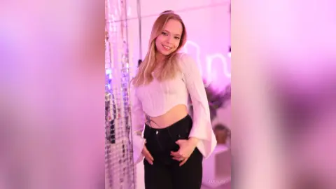 LucyLoy smiles while posing in a white crop top and black pants in a pink lit room.