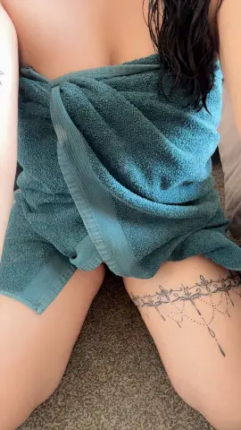 Woman with thigh tattoo wearing a teal towel wrap sitting on a carpeted floor in a relaxed mood.