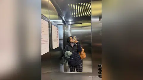 Young woman in a black puffer jacket taking a mirror selfie in a modern metal elevator.