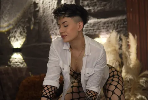 Young woman with short dark hair in a white shirt and fishnet tights posing in a moody, dimly lit room.