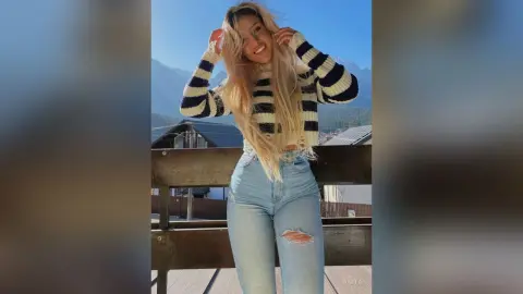 Anita smiles while wearing a black and white striped long-sleeve crop top and light blue ripped denim jeans outdoors.