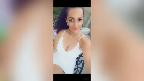 AishaApril wearing a white tank top or bodysuit, looking at the camera with her dark curly hair down outdoors.