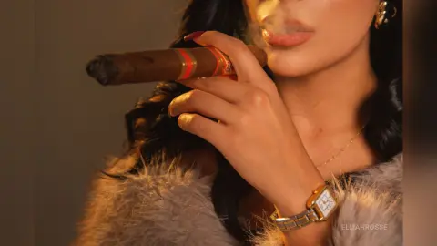Elegant woman in a grey faux fur coat smoking a large cigar in warm, moody lighting.