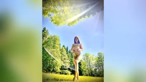 EmaLowee stands in a sunny grassy field surrounded by trees, wearing a light-colored bikini that shows off her bare stomach and legs.