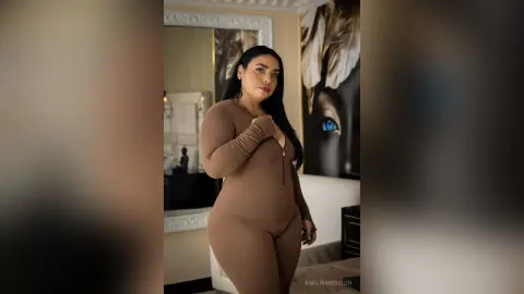 Curvy woman in a brown long-sleeve bodysuit posing confidently in a modern, stylishly lit bedroom.