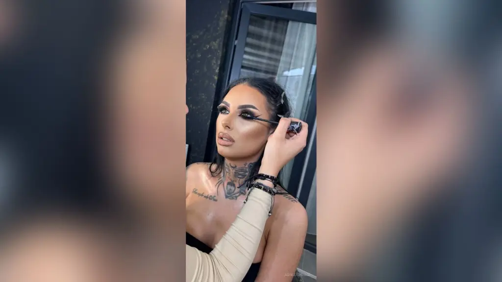 AshleighPage — Glamorous woman with dramatic winged eyeliner and tattoos applying mascara in a bright indoor setting.
