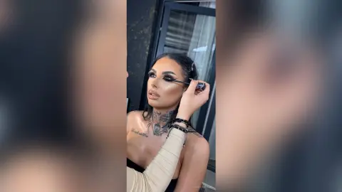 Glamorous woman with dramatic winged eyeliner and tattoos applying mascara in a bright indoor setting.