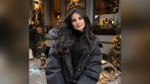 Smiling woman in a black puffer jacket and turtleneck sweater standing by festive Christmas trees outdoors.