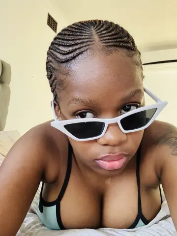 Young woman with cornrow braids and white sunglasses relaxing in a bedroom setting.