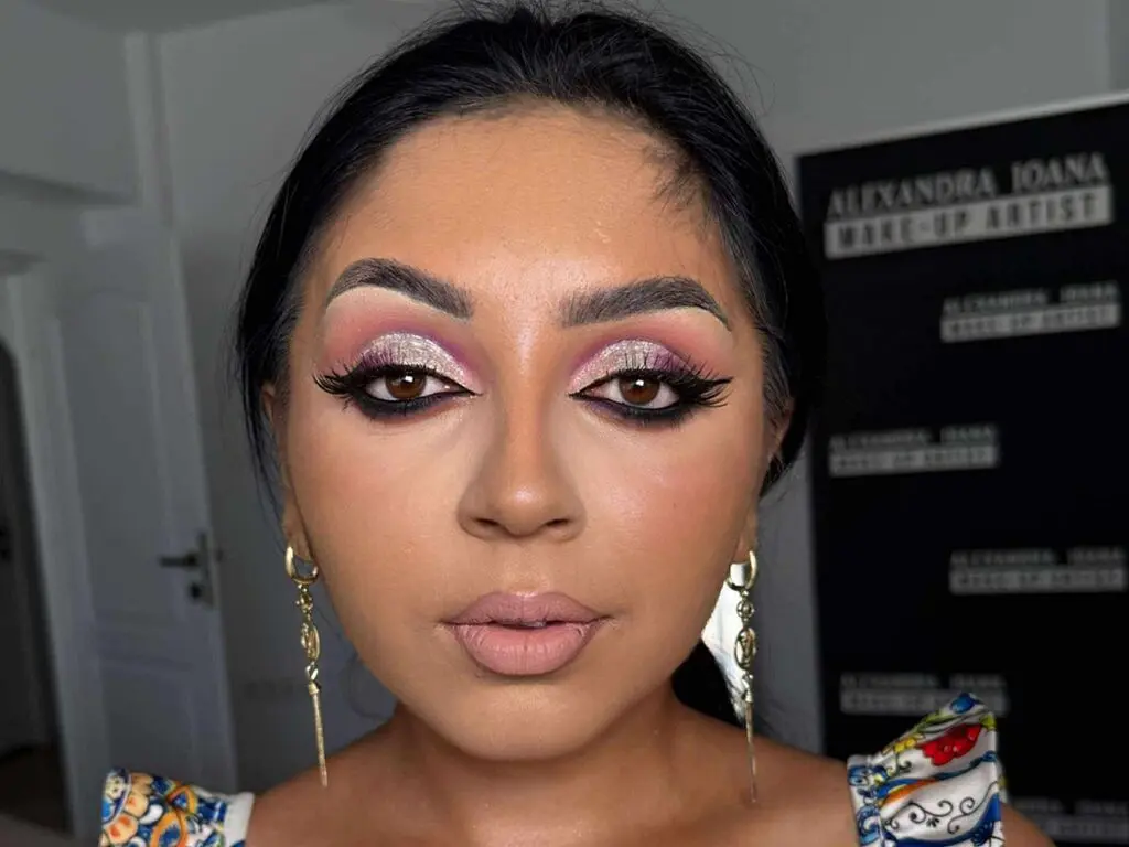 Close-up of a woman with glamorous pink glitter eye makeup and gold earrings in a professional studio setting.