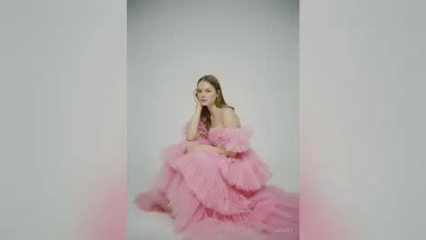 Elegant woman in a voluminous pink ruffled tulle gown posing against a minimalist grey background.