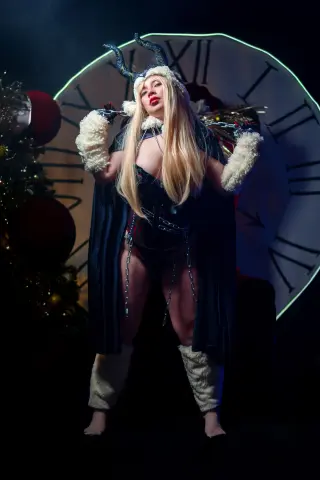Nyxaria poses in a dark fantasy outfit with horns and a cape, standing in front of a large clock face.
