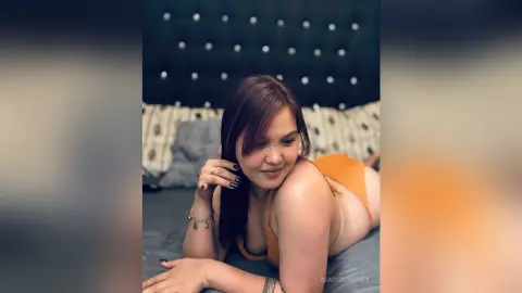 Smiling woman in an orange top relaxing on a bed in a cozy, moody bedroom setting.